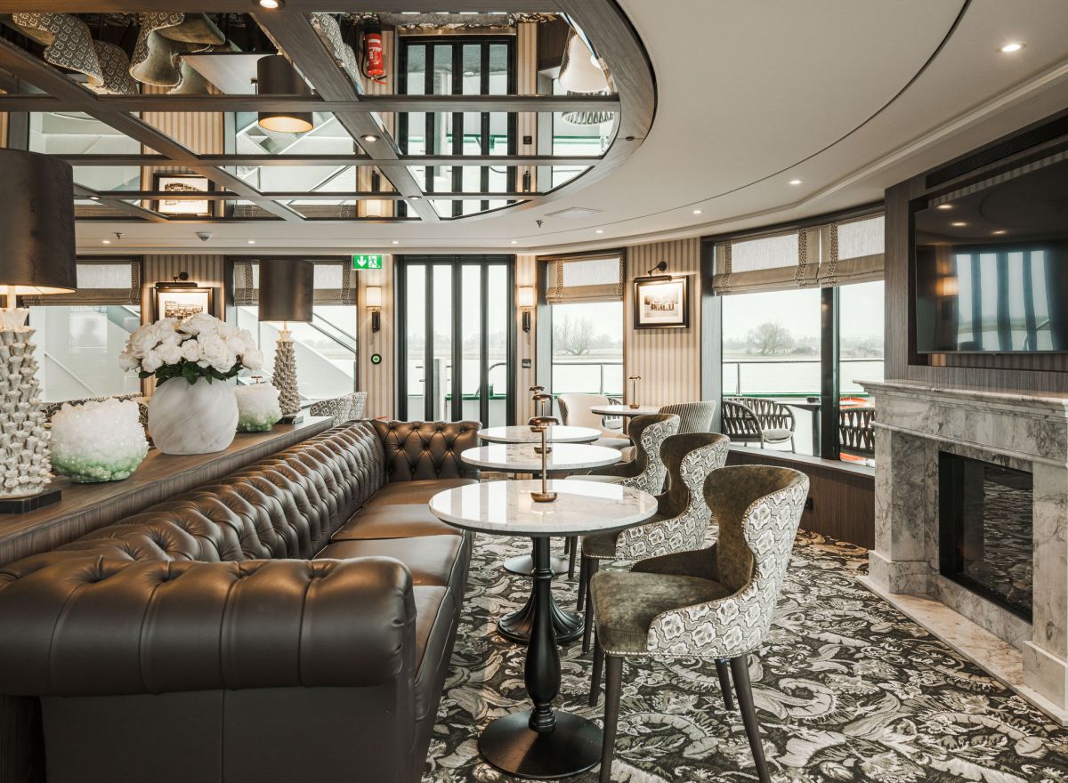 Refined onboard dining lounge with blue velvet chairs, patterned upholstery, and large riverside windows.